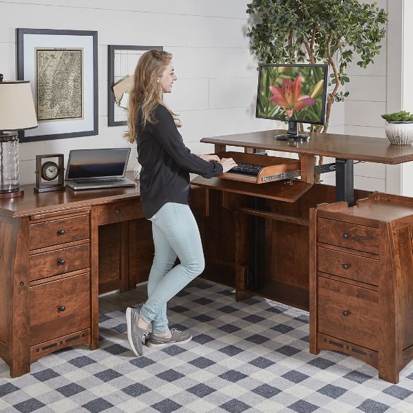 Traditional style Boulder Creek L-shaped desk with wood finish, featuring a sit-to-stand adjustable drawer on the left and a women standing and going on her computer