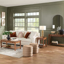 A living room scene featuring furniture pieces including a white sofa with pillows, a brown end table, a wooden coffee table, and a sofa table, all arranged in a stylishly decorated room with green walls and wooden flooring.