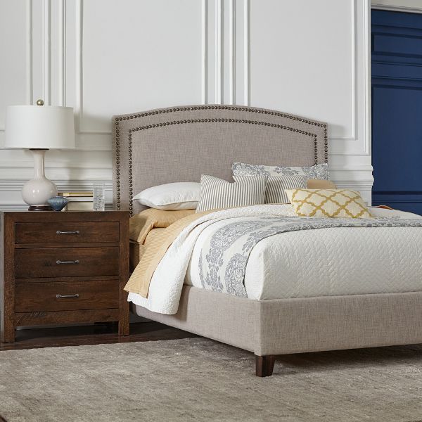 The image features the Gabrielle Upholstered Bed with a solid oak hardwood frame and a black nickel nailhead upholstery. The bed is dressed with a variety of pillows and is positioned in a bedroom setting with a decorative lamp and a nightstand.