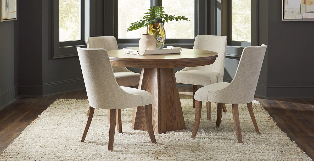 Brogan round pedestal dining table in a natural wood finish, paired with four cream upholstered chairs in a contemporary dining room with dark walls, large windows, and neutral decor.