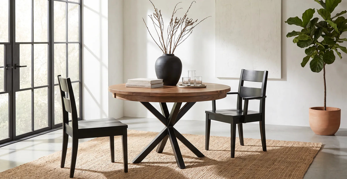Brooklyn Round Pedestal Dining Table with a warm wood top and black X-shaped base, paired with black Carson Dining Chairs in a bright, modern dining room with large windows.