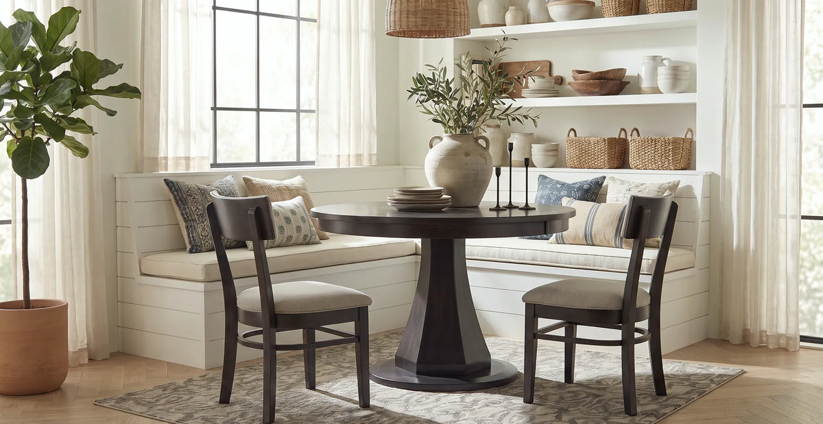 Emerson round pedestal dining table in a rich dark wood finish, paired with upholstered chairs and a cushioned corner bench in a bright, cozy dining nook with natural decor.