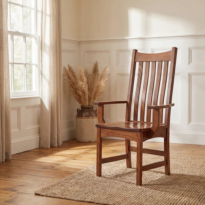 A wooden armchair with a tall slatted back and armrests, placed on a woven rug in a bright room with white paneled walls, a window, and a vase of dried grass.