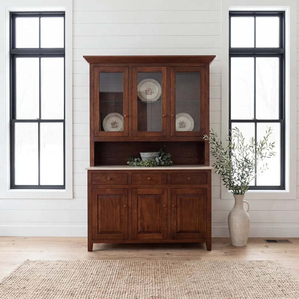A dark brown wooden hutch with glass-front upper cabinets, a stone-look countertop, and three lower cabinet doors, staged in a bright room with white shiplap walls, black-framed windows, a woven rug, and a tall vase with greenery.