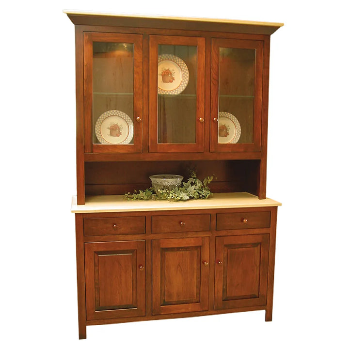 A mid-century wooden hutch and sideboard cabinet with a warm medium-brown finish, featuring three upper glass-front doors with decorative plates, a central open shelf with greenery and a bowl, and three lower cabinet doors with drawers above. The cabinet has a light beige stone or laminate top and bottom surfaces.