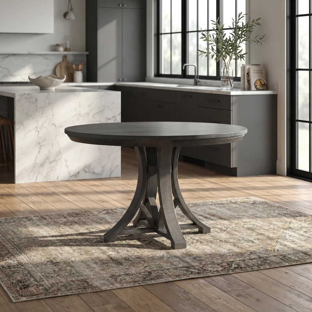 A round dark gray wooden dining table with a sculptural pedestal base, set on a distressed beige and gray area rug in a modern kitchen with wood floors.