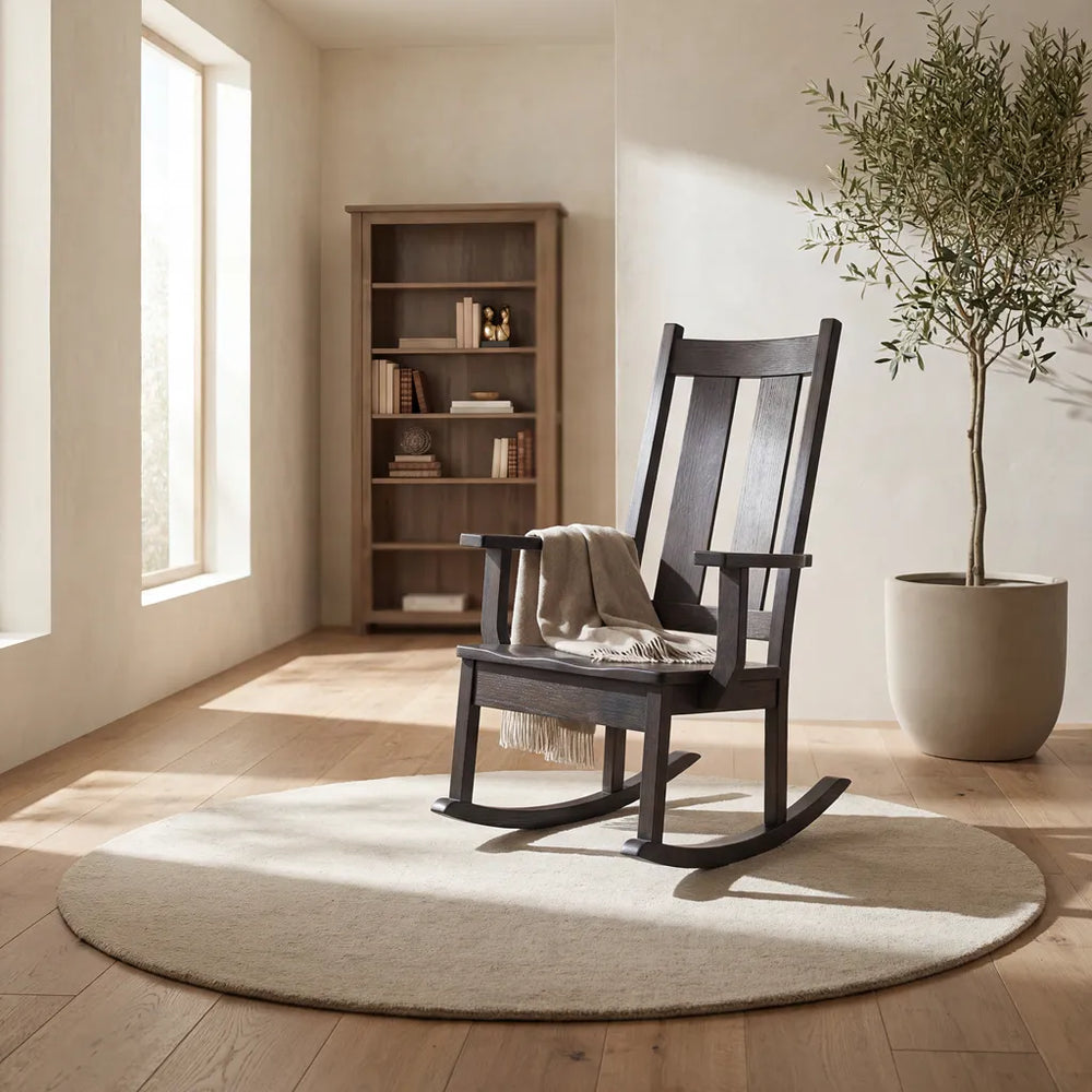 A dark wood rocking chair with a beige throw blanket sits on a round light beige rug in a sunlit room with light wood floors, a tall bookshelf, and a potted tree.
