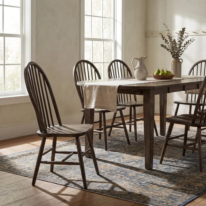 A dark wood dining table with matching spindle-back chairs in a bright, sunlit dining room. The table is set with a beige table runner, a white ceramic pitcher, and a bowl of green apples, all arranged on a blue and beige patterned area rug over a hardwood floor.