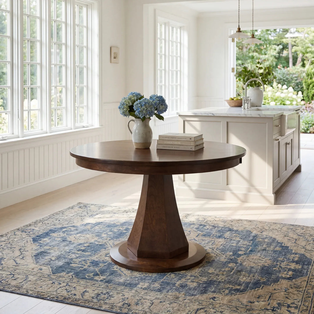 A round dark wood pedestal table with a ceramic vase of blue hydrangeas and a stack of books on top, set on a blue and beige patterned area rug in a bright, modern dining room.