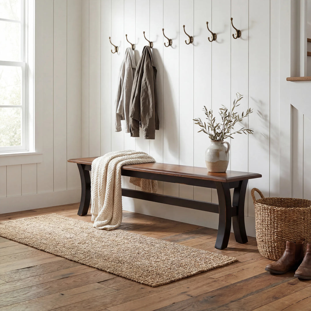 A rustic wooden bench with dark legs sits in a bright entryway with white shiplap walls, wall hooks, a cream throw blanket, a vase with branches, a woven basket, and boots.