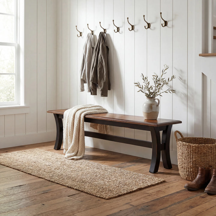 A rustic wooden bench with dark legs sits in a bright entryway with white shiplap walls, wall hooks, a cream throw blanket, a vase with branches, a woven basket, and boots.