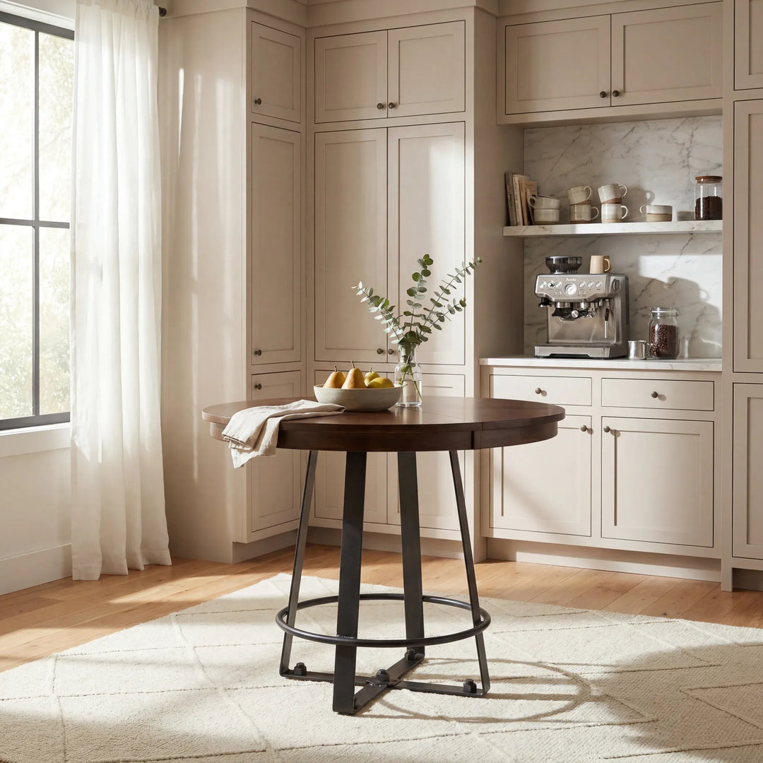 A round dark wood bar-height table with black metal legs stands in a bright, sunlit kitchen or dining area with cream cabinetry, a marble backsplash, and a vase of greenery on top.