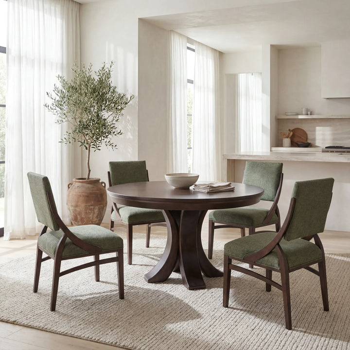 A round dark wood dining table with four upholstered chairs in sage green, arranged on a light woven rug in a bright, modern dining room.