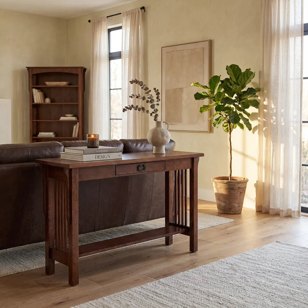 A dark brown wooden console table in a bright, modern living room setting with a leather sofa, bookshelf, and potted plant.
