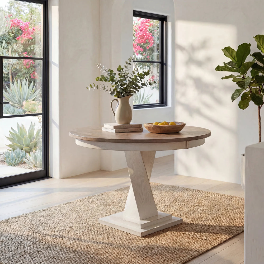Round wooden table with a white pedestal base in a bright room with large windows and plants.