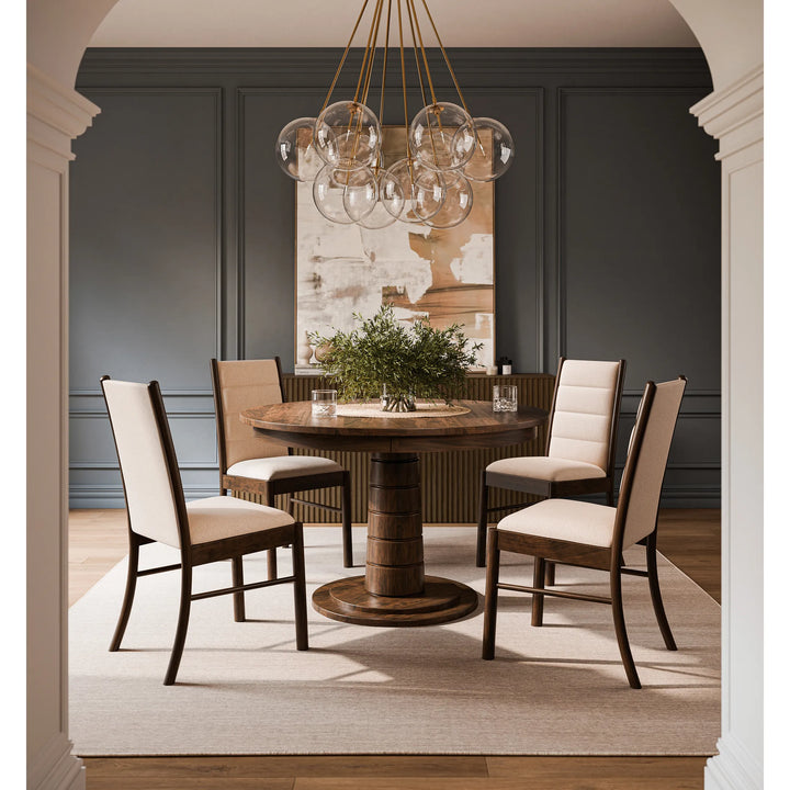 A round wooden dining table with four upholstered chairs in a staged dining room. The room features dark gray paneled walls, a large glass bubble chandelier, and a light beige rug.