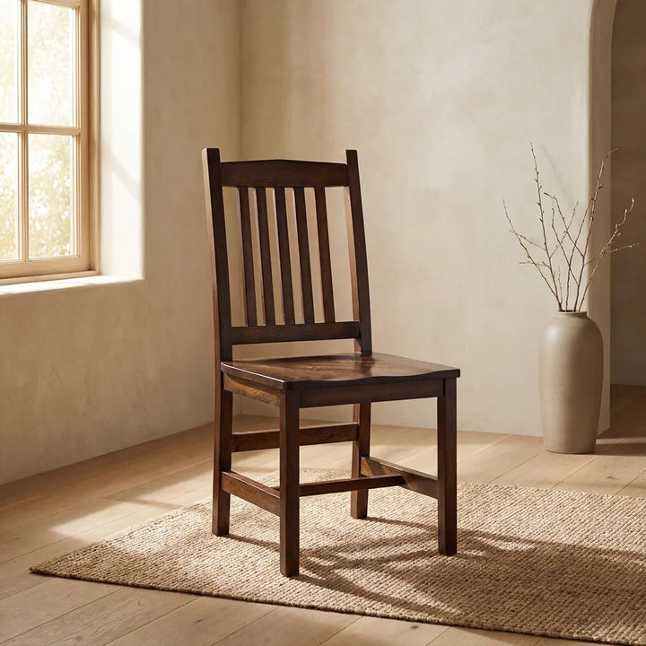 A dark brown wooden dining chair with a slatted backrest, positioned on a woven jute rug in a warm, sunlit room with beige walls and a large ceramic vase in the background.