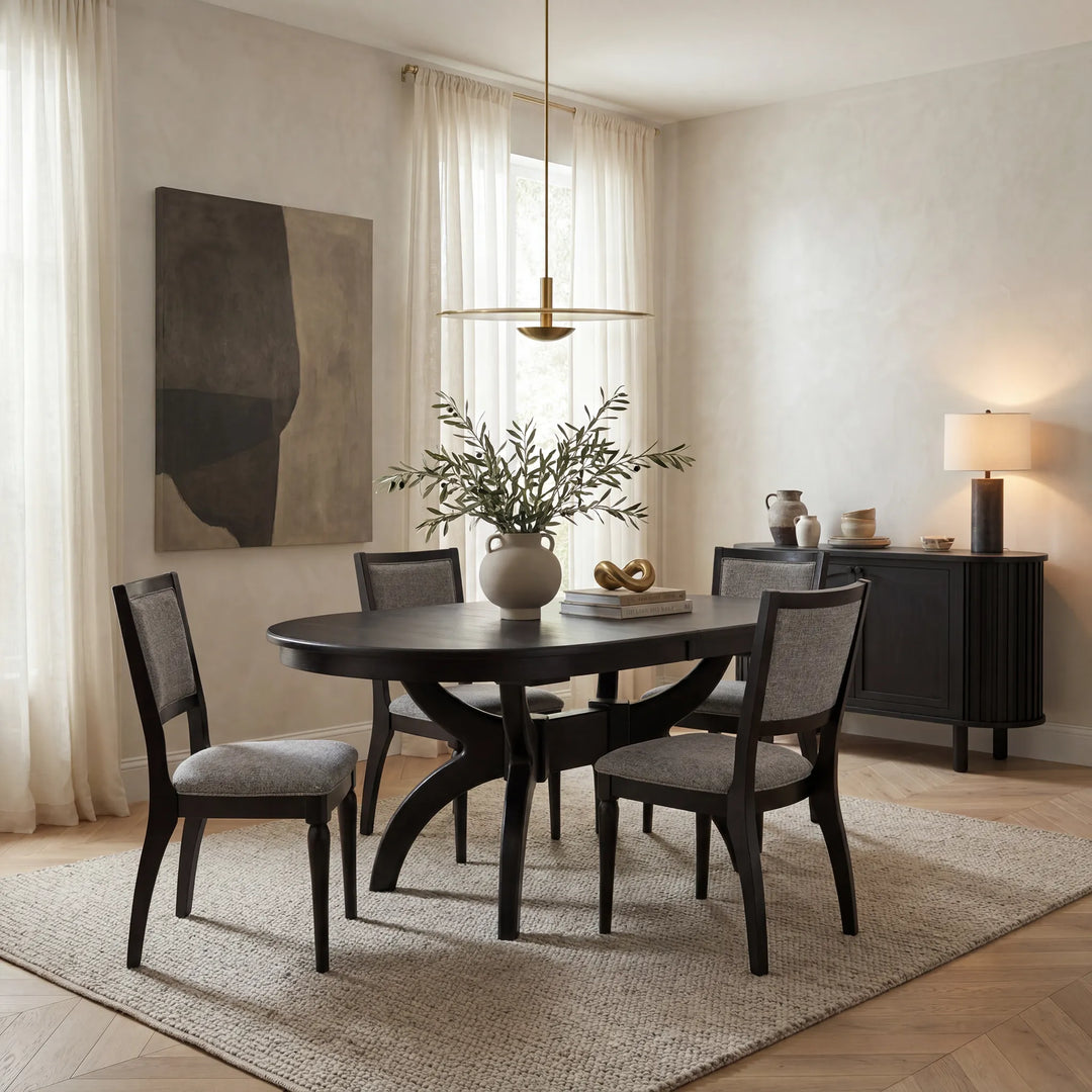 A dark round dining table with four upholstered chairs in a styled dining room. The room features neutral beige walls, a large abstract painting, a hanging brass pendant light, and a sideboard with a lit lamp in the background.
