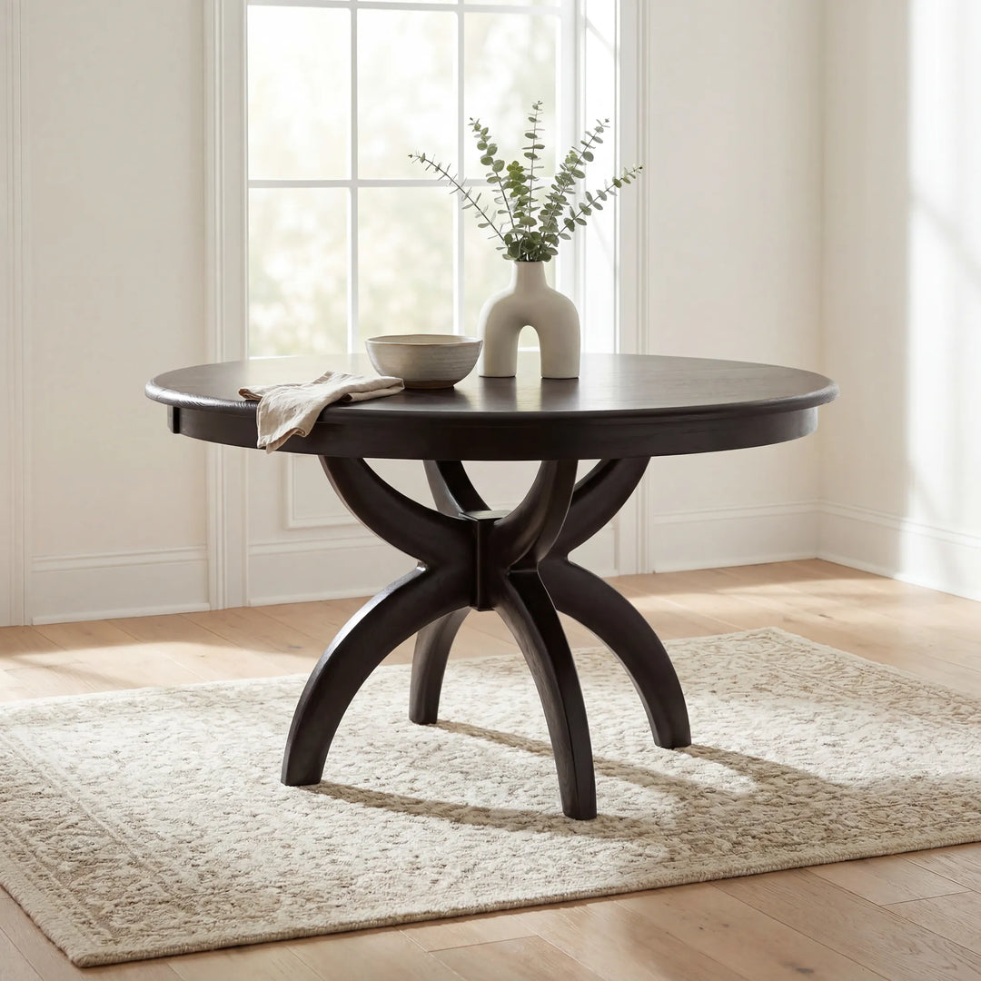 A round dark wood dining table with a sculptural multi-leg base, styled with a ceramic vase and bowl on a light rug in a bright room.