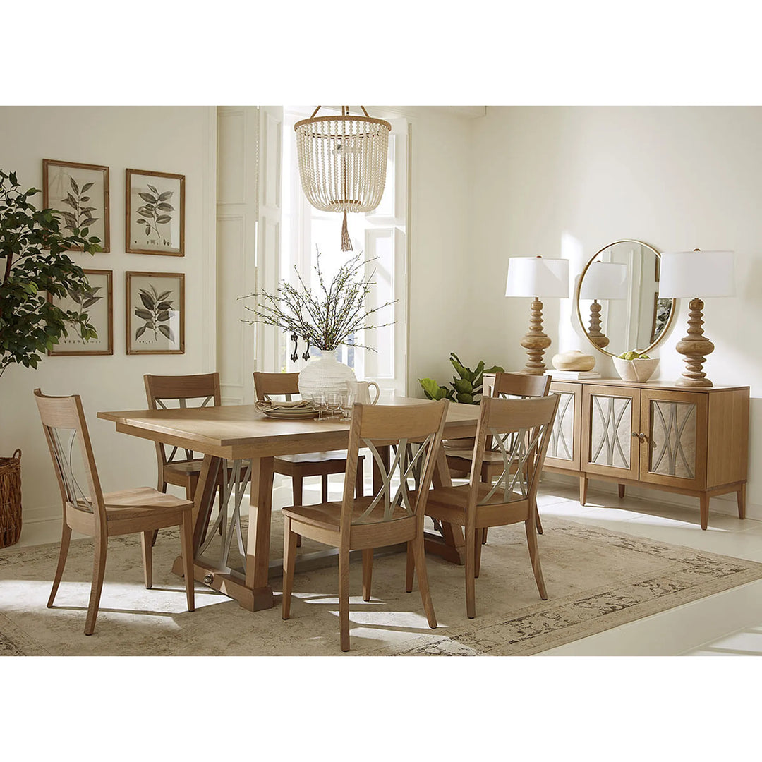 A light oak dining room set featuring a rectangular wooden table and six matching chairs with woven backrests, styled in a bright, modern dining room with a sideboard, lamps, and framed botanical prints.
