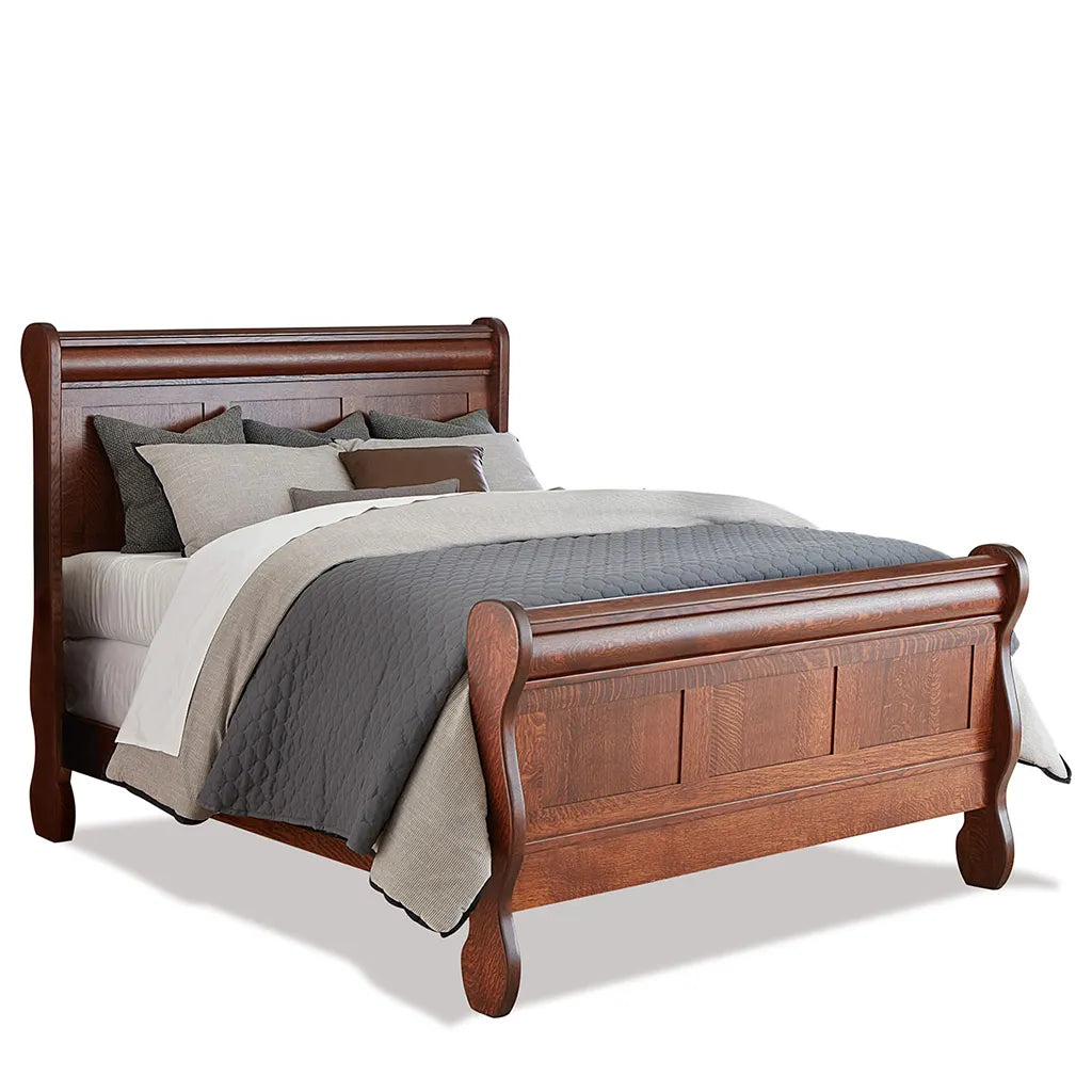 A classic wooden sleigh bed with a warm medium-brown finish, styled with gray and beige bedding, set against a plain white background.