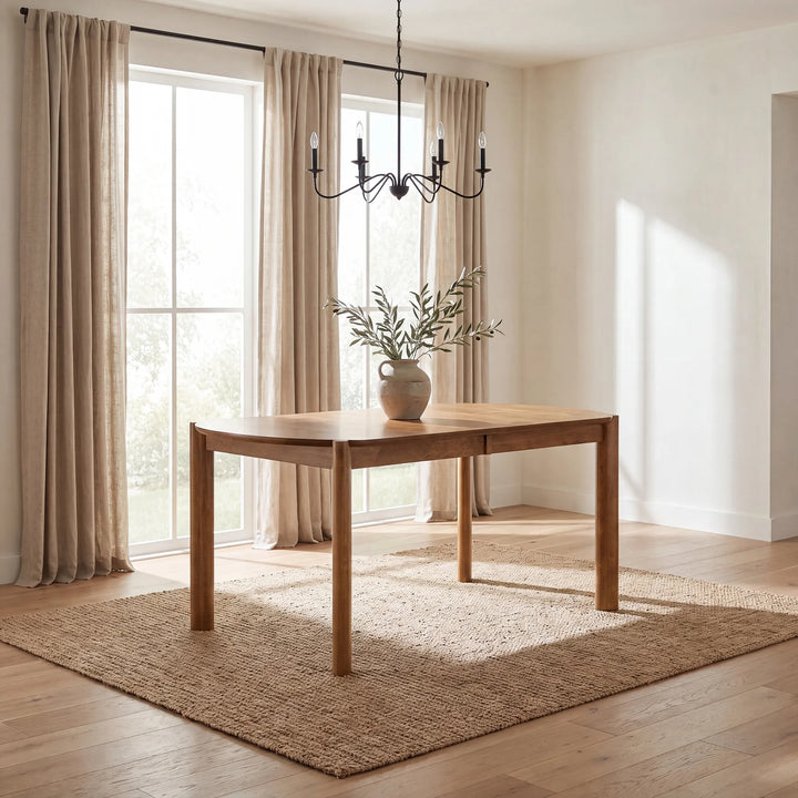 A rectangular wooden dining table with a light beige ceramic vase of greenery on top, set on a woven jute rug in a bright, modern dining room with beige curtains and wood floors.