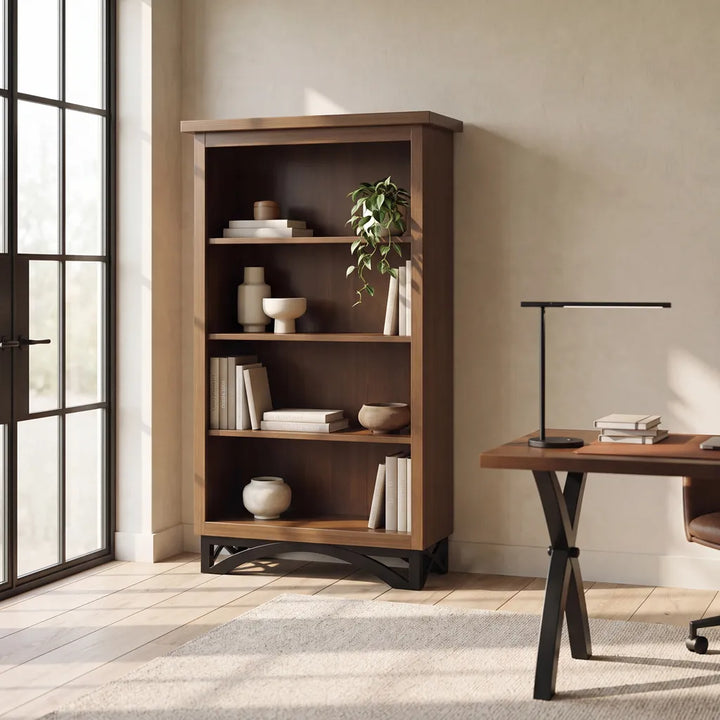 A tall wooden bookcase with five shelves stands in a bright room next to a window, styled with books, vases, bowls, and a small potted plant. A wooden desk with a black lamp is visible on the right.
