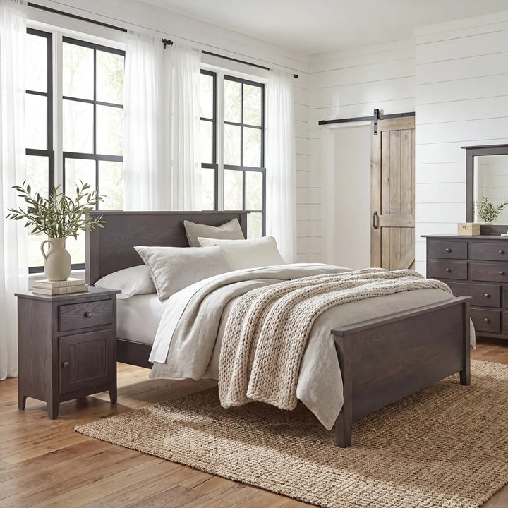 A rustic bedroom scene featuring a dark wood bed frame with white and beige bedding, a matching dark wood nightstand, a woven jute rug, and a barn-style sliding door in the background.