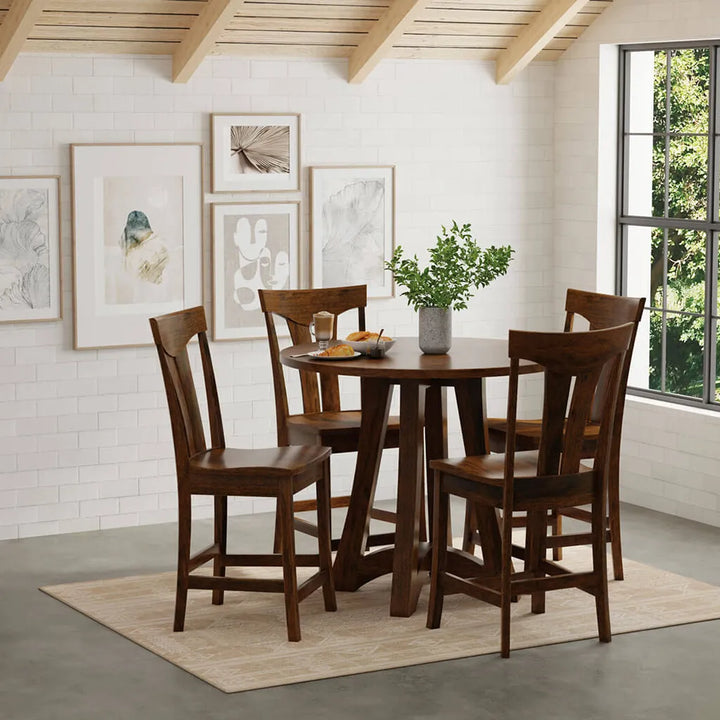 A 5-piece counter height dining set featuring a round dark brown wood table and four matching bar-height chairs, styled in a bright dining room with a white brick wall, framed art, and a window.