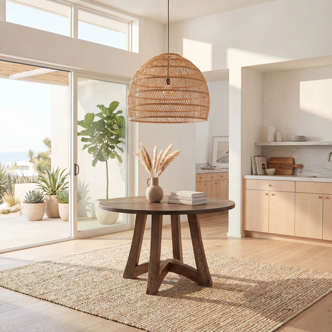 A round dark wood dining table with three angled legs, styled in a bright, modern coastal kitchen with a woven rattan pendant light overhead and a natural jute rug underneath.