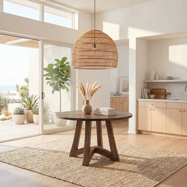 A round dark wood dining table with three angled legs, styled in a bright, modern coastal kitchen with a woven rattan pendant light overhead and a natural jute rug underneath.
