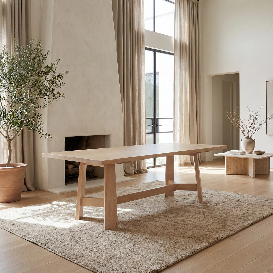 A light natural wood dining table with a rectangular top and tapered legs, styled in a bright, modern minimalist room with beige walls, a fireplace, and a shag rug.