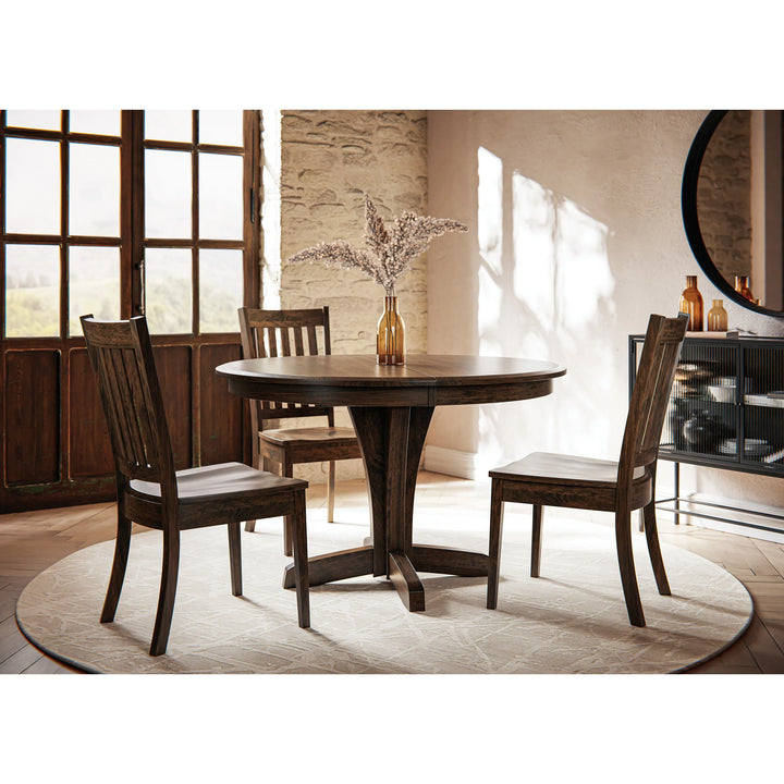 A round dark wood dining table with four matching wooden chairs, styled in a rustic dining room setting with a stone wall, large windows, and a round rug.