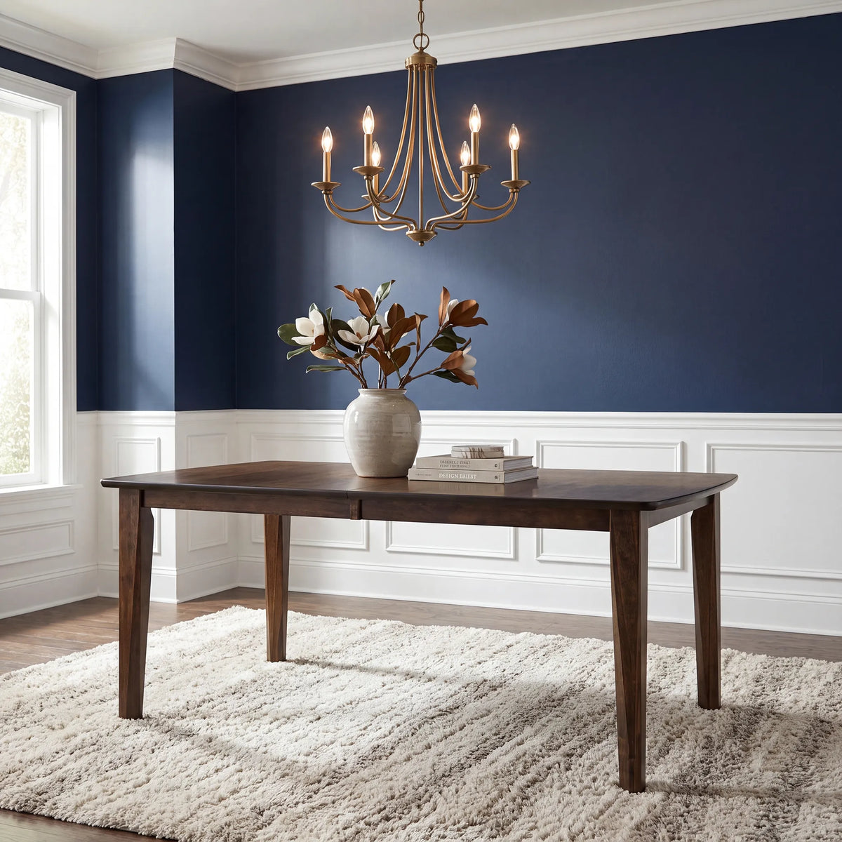A dark wood rectangular dining table in a styled room with a deep blue wall, white wainscoting, a hanging brass chandelier, a beige rug, and a vase of flowers on top.